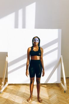 Full body confident African American female with braided long curly hairstyle wearing sport bra and leggings standing against white backdrop in minimal interior room with white wall and wooden parquet floor and looking at camera