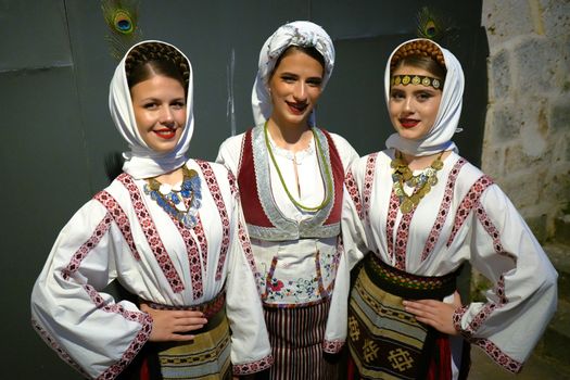 Three women in traditional Balkan attire with colorful designs, smiling indoors.