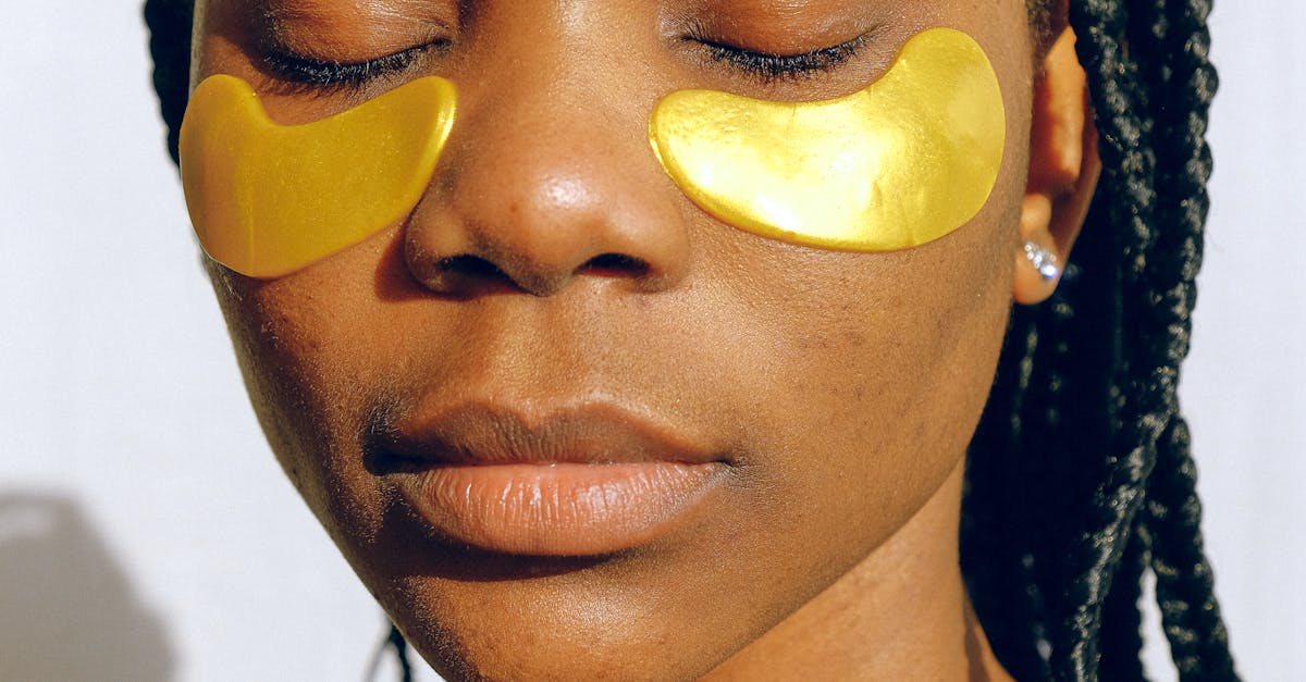Closeup of young calm African American female relaxing with eyes closed applying eye patches against dark circles and wrinkles during cosmetic procedure of skin care on white wall background