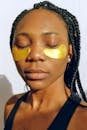 Closeup of young calm African American female relaxing with eyes closed applying eye patches against dark circles and wrinkles during cosmetic procedure of skin care on white wall background