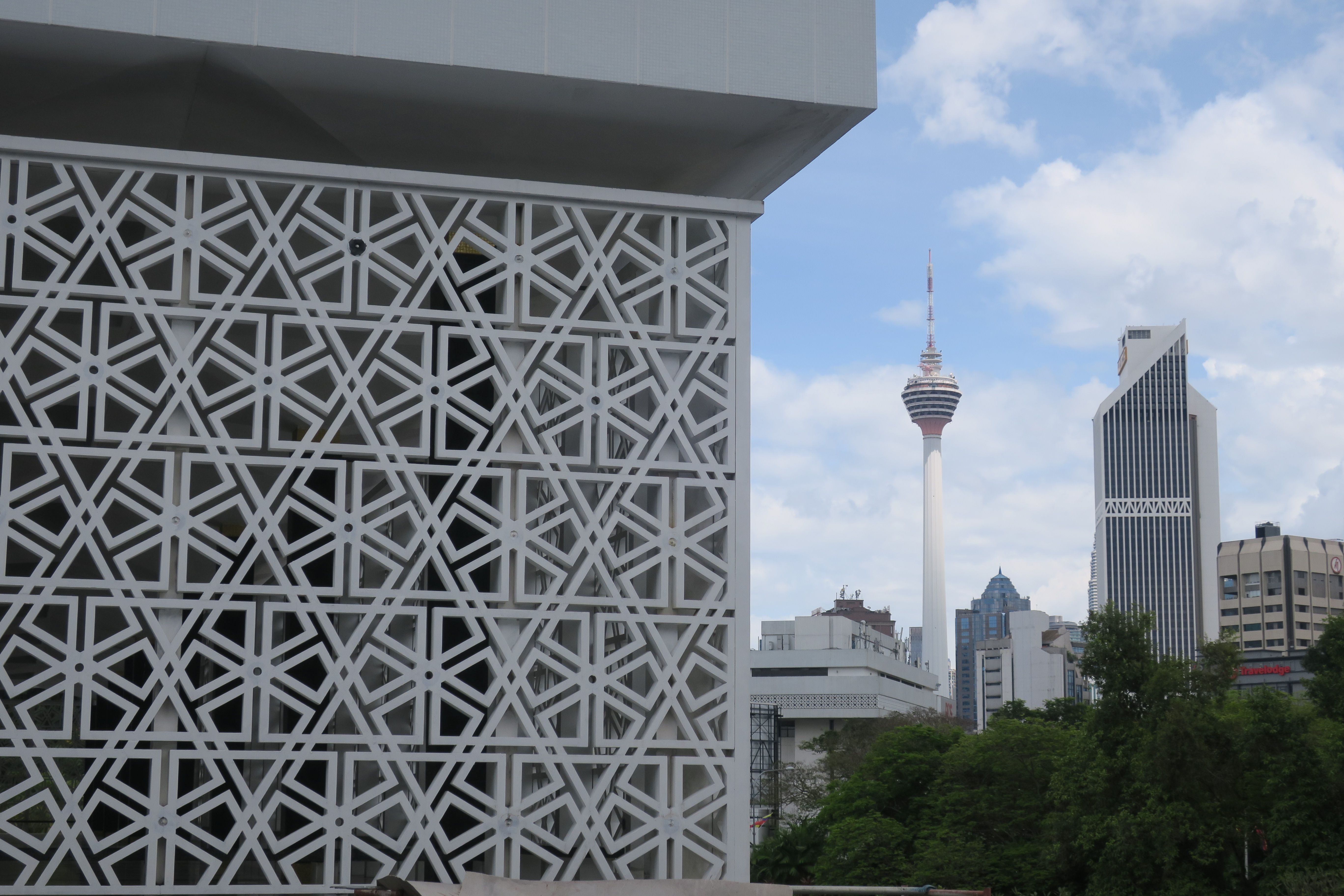 A view of Kuala Lumpur Tower amidst modern buildings and intricate architecture, under a clear sky.