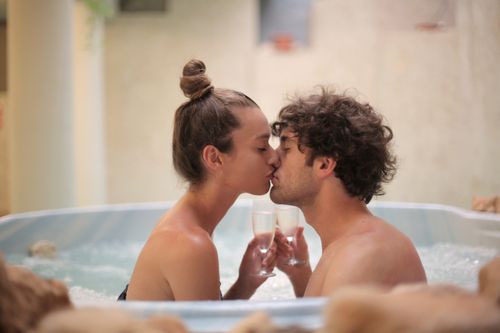 Free Side view of young tender man and woman sitting in bathtub with drinks in wine glasses and kissing each other enjoying weekend while spending time together in spa center Stock Photo