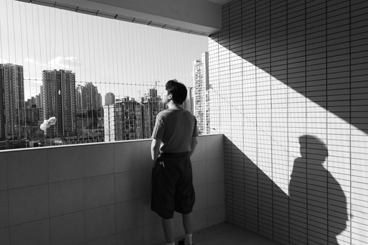 Person in shadow gazes at city skyline from balcony, creating a reflective mood.
