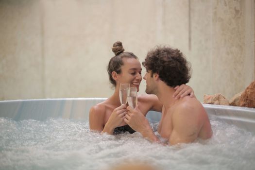 Cheerful young man and woman enjoying time together sitting in round bath with water babbles while drinking champagne and looking at each other with love in spa center during weekend