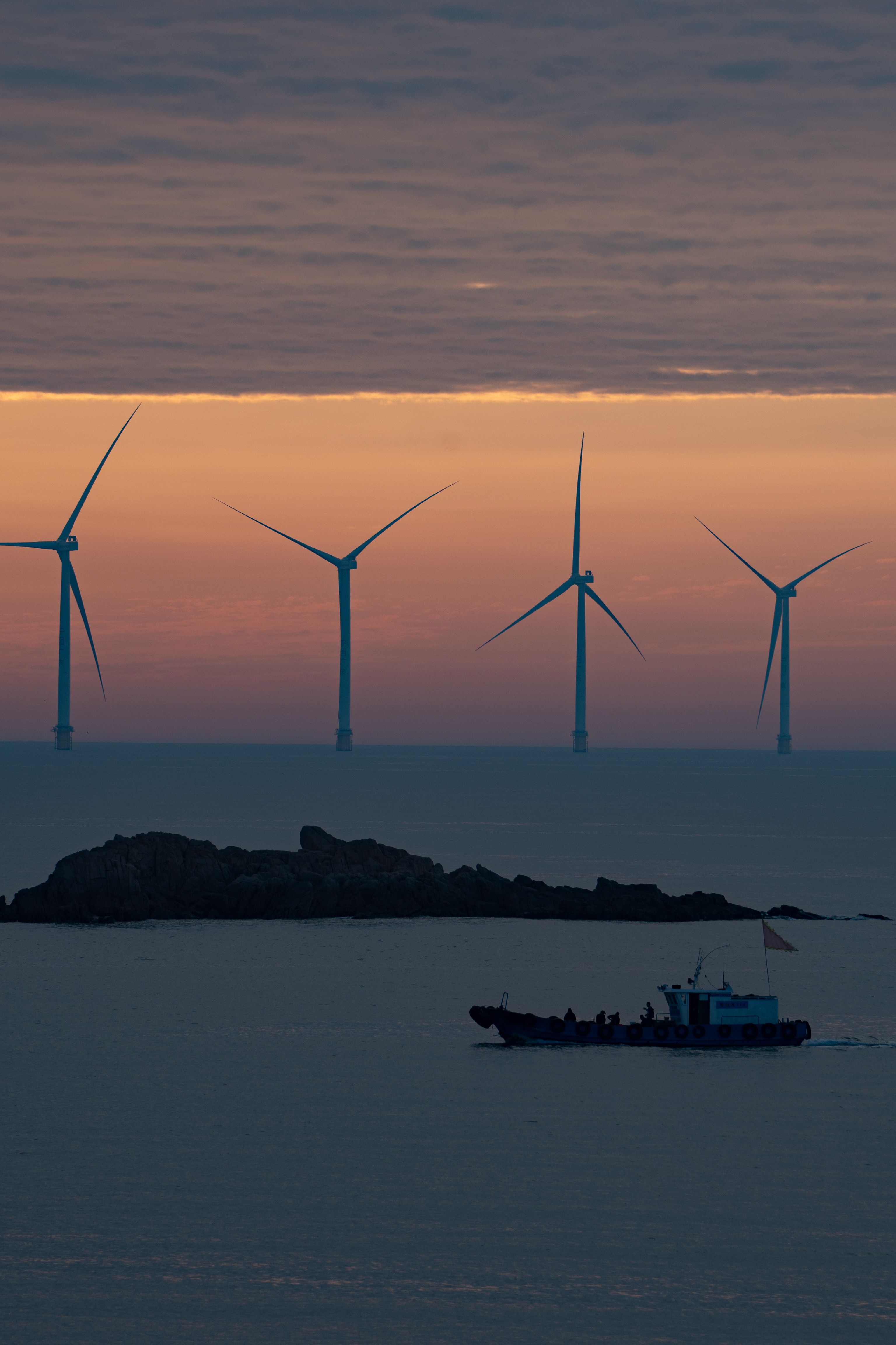Silhouetted wind turbines and a boat against a vibrant sunset over the sea in Ningbo, China.