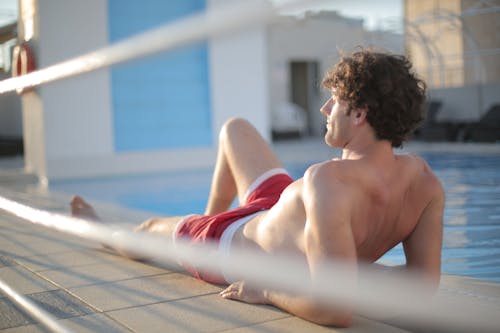 Side view of shirtless male in red shorts chilling on concrete border near pool while lying on lower back resting on hands