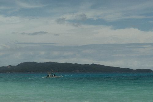 Free A small boat sails on turquoise waters against a backdrop of a lush green island under a cloudy sky. Stock Photo