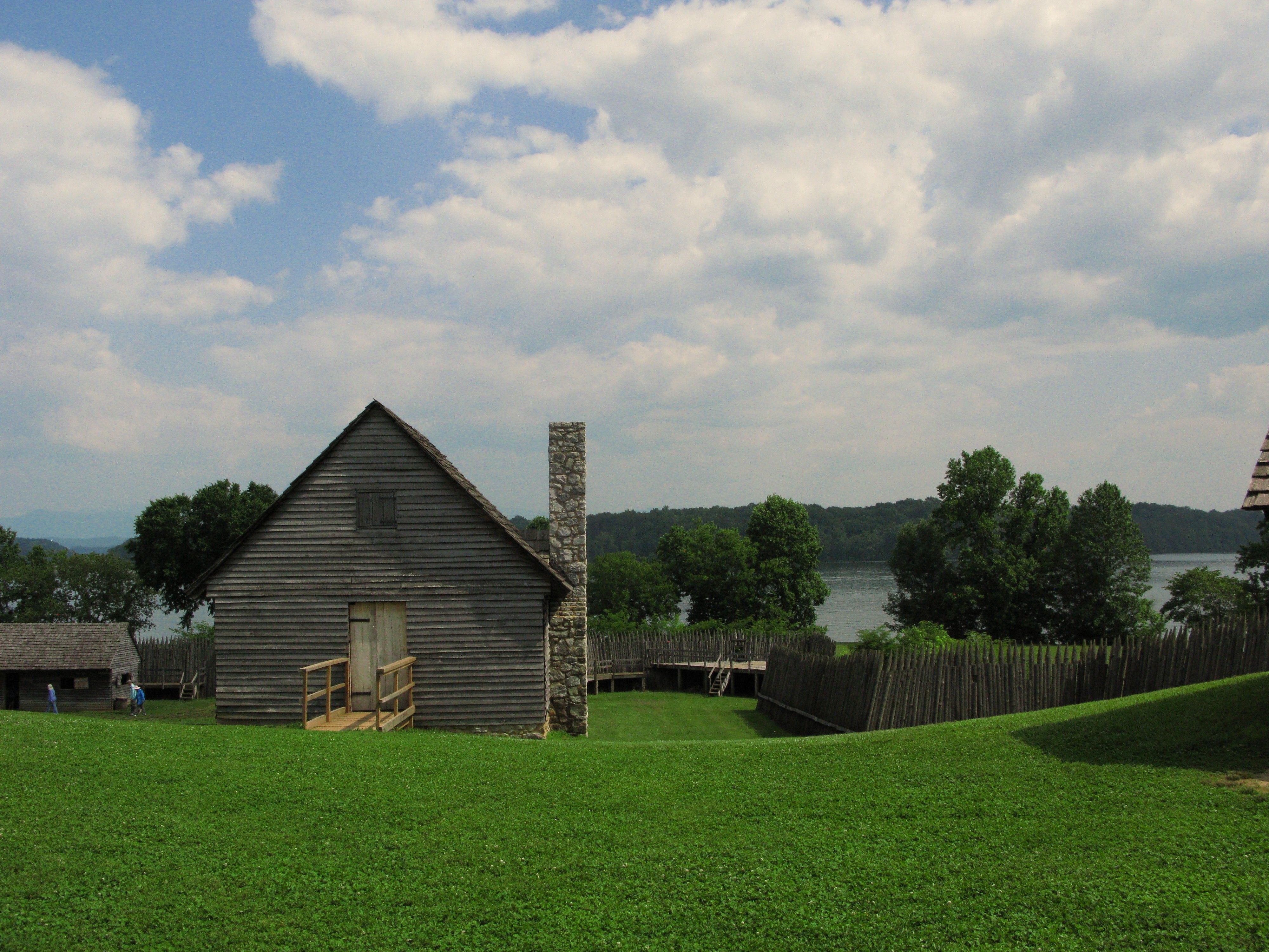 A rustic wooden cabin and picket fence by a tranquil lake under a bright summer sky.