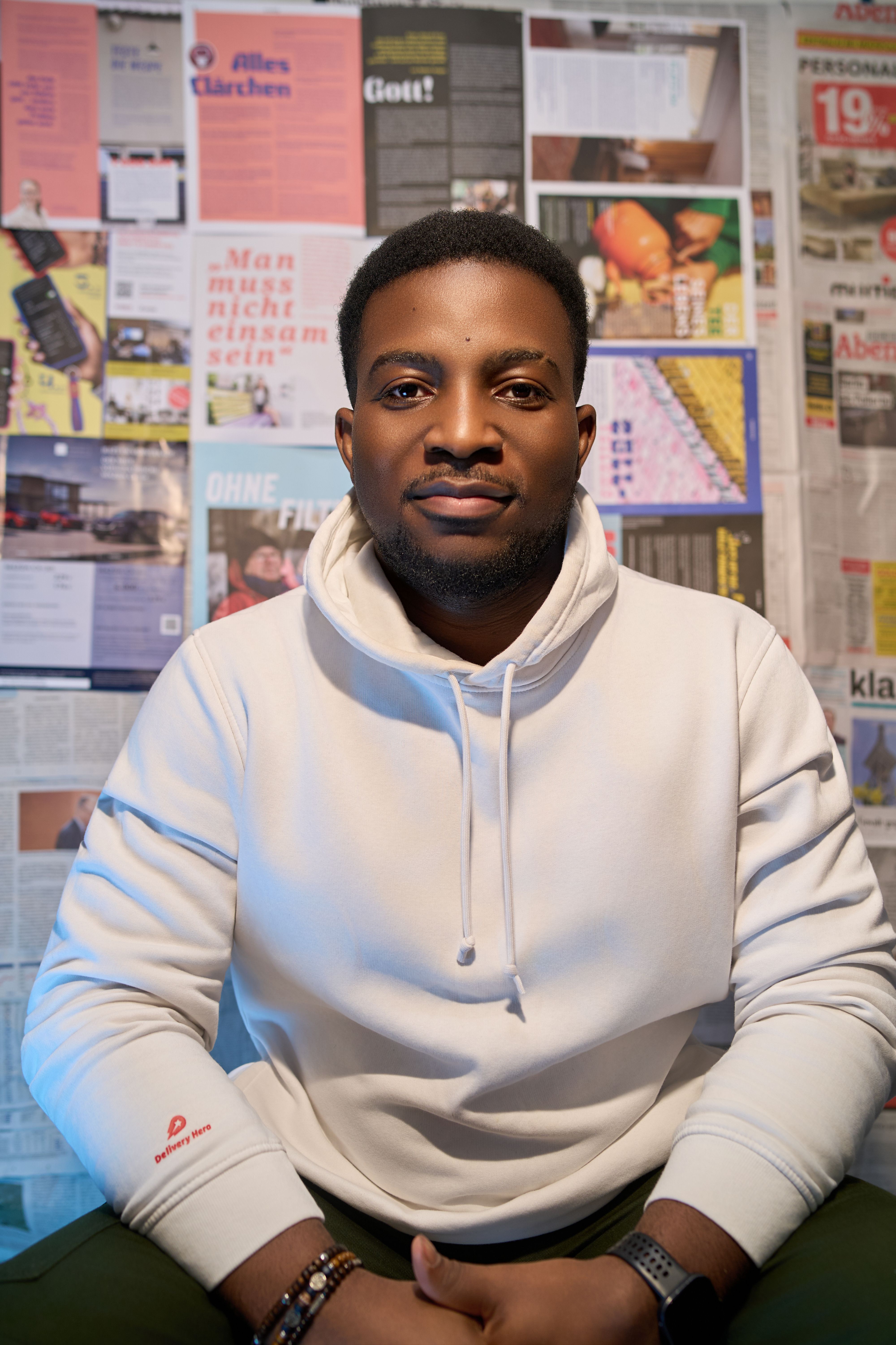Man in white hoodie seated against a newspaper collage backdrop.