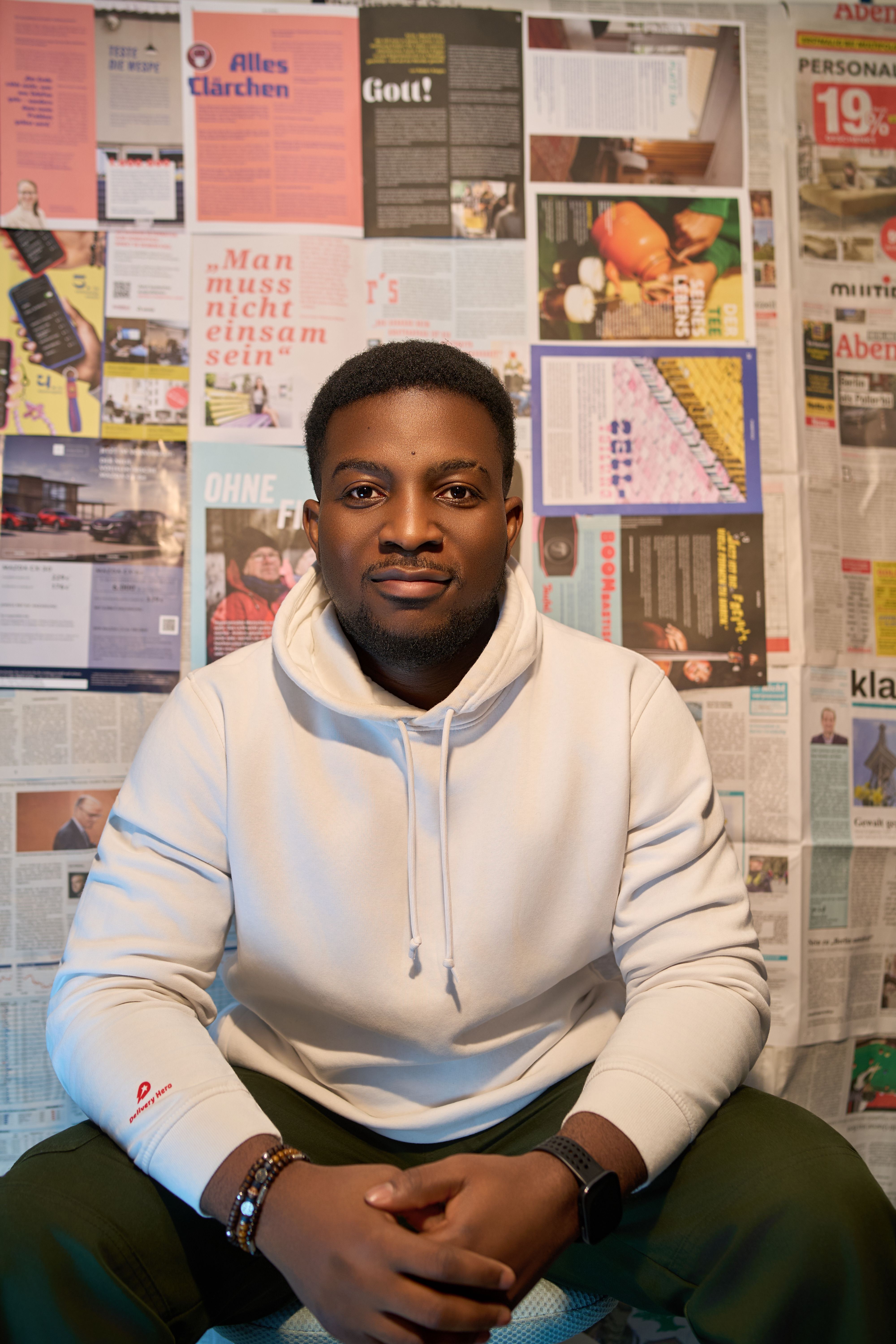 A confident man in a white hoodie poses against a wall covered with newspapers.