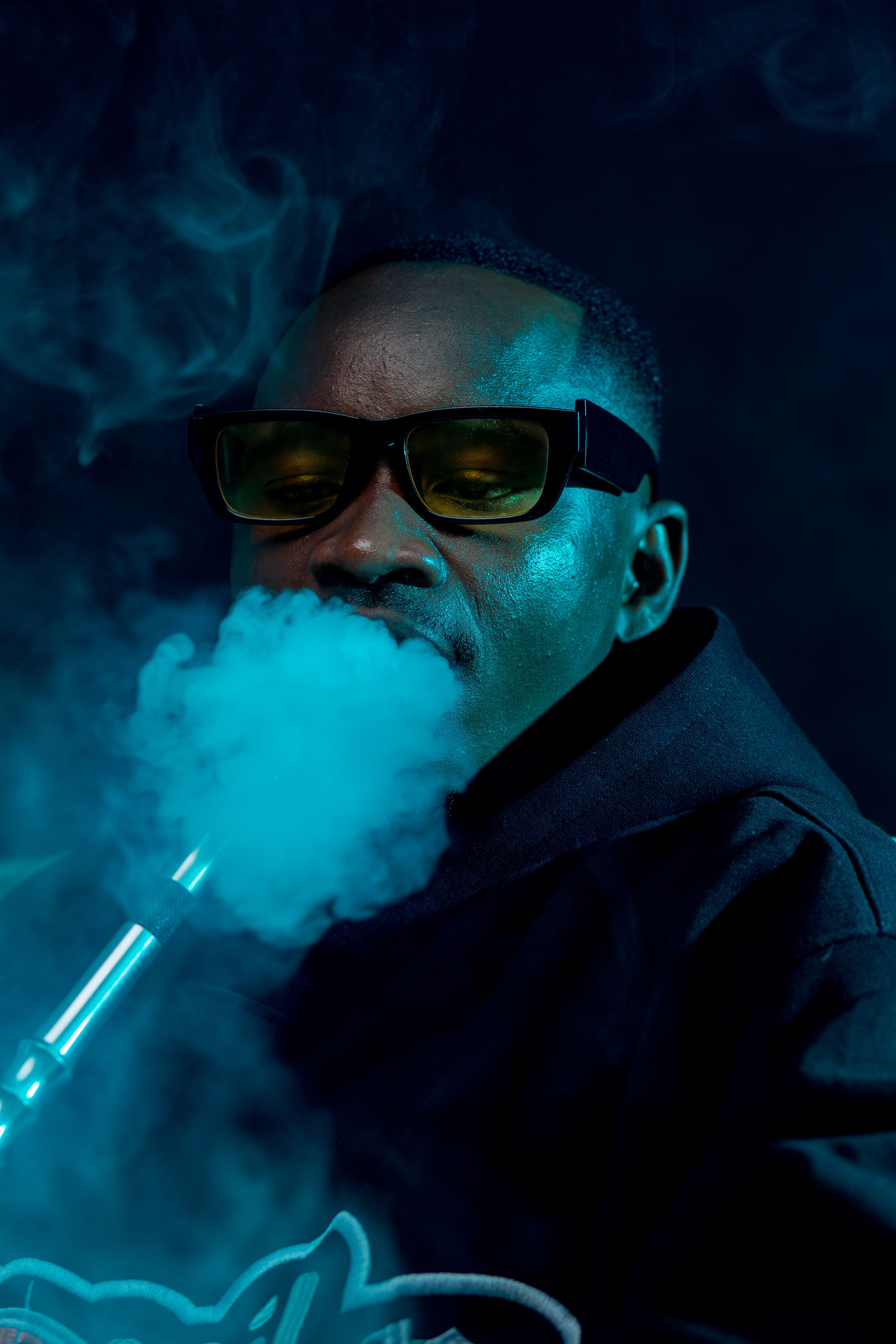A man wearing sunglasses smokes a hookah against a dark background.