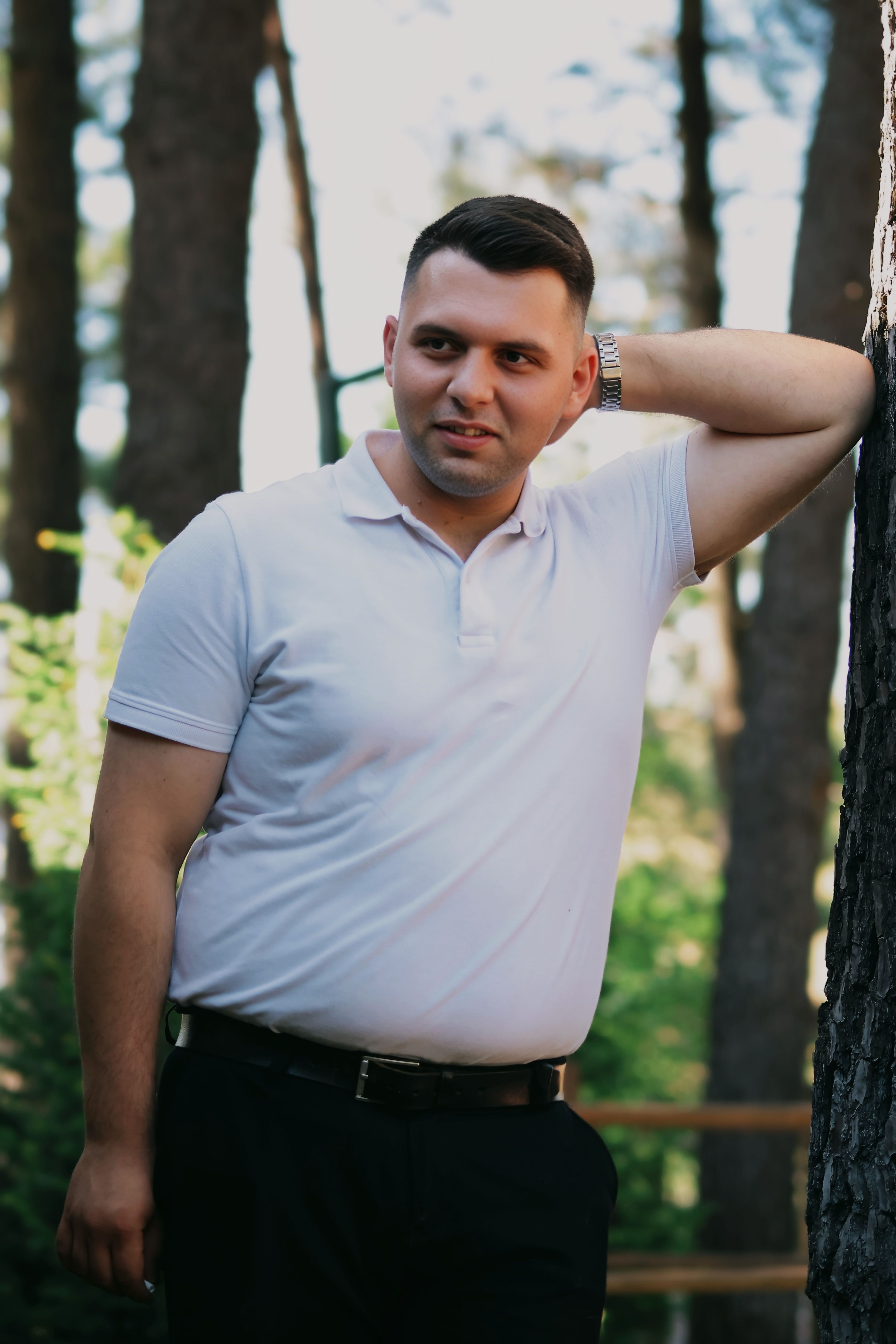 A man in a white shirt stands casually in a forest, leaning against a tree.