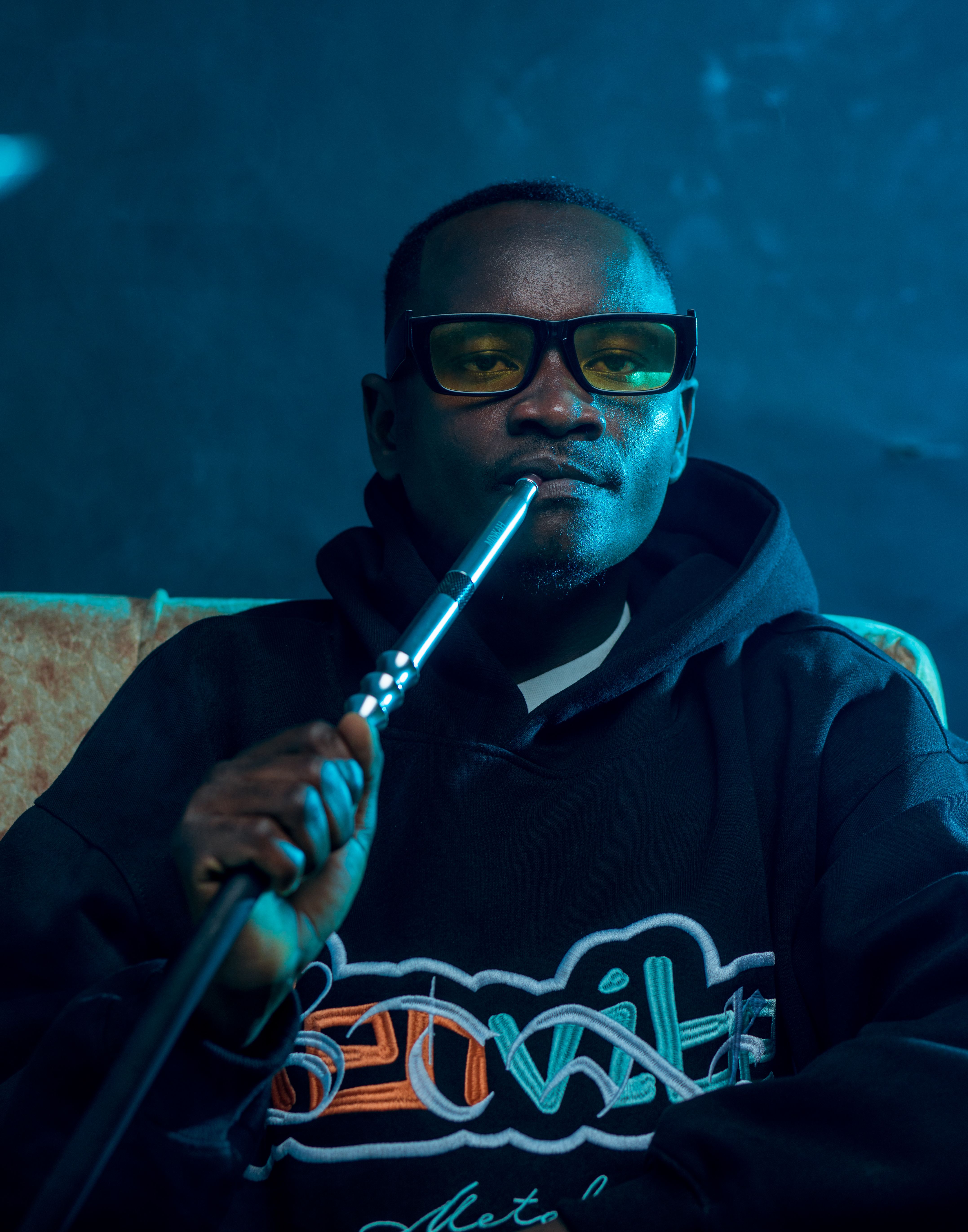 A man wears sunglasses and a hoodie while relaxing indoors with a hookah pipe.