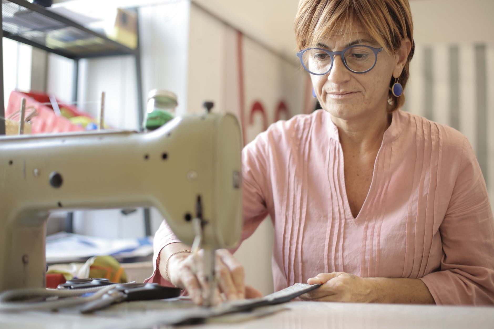 Focused mature female in casual clothes and eyeglasses sitting at table and sewing leather purse while working in modern workshop