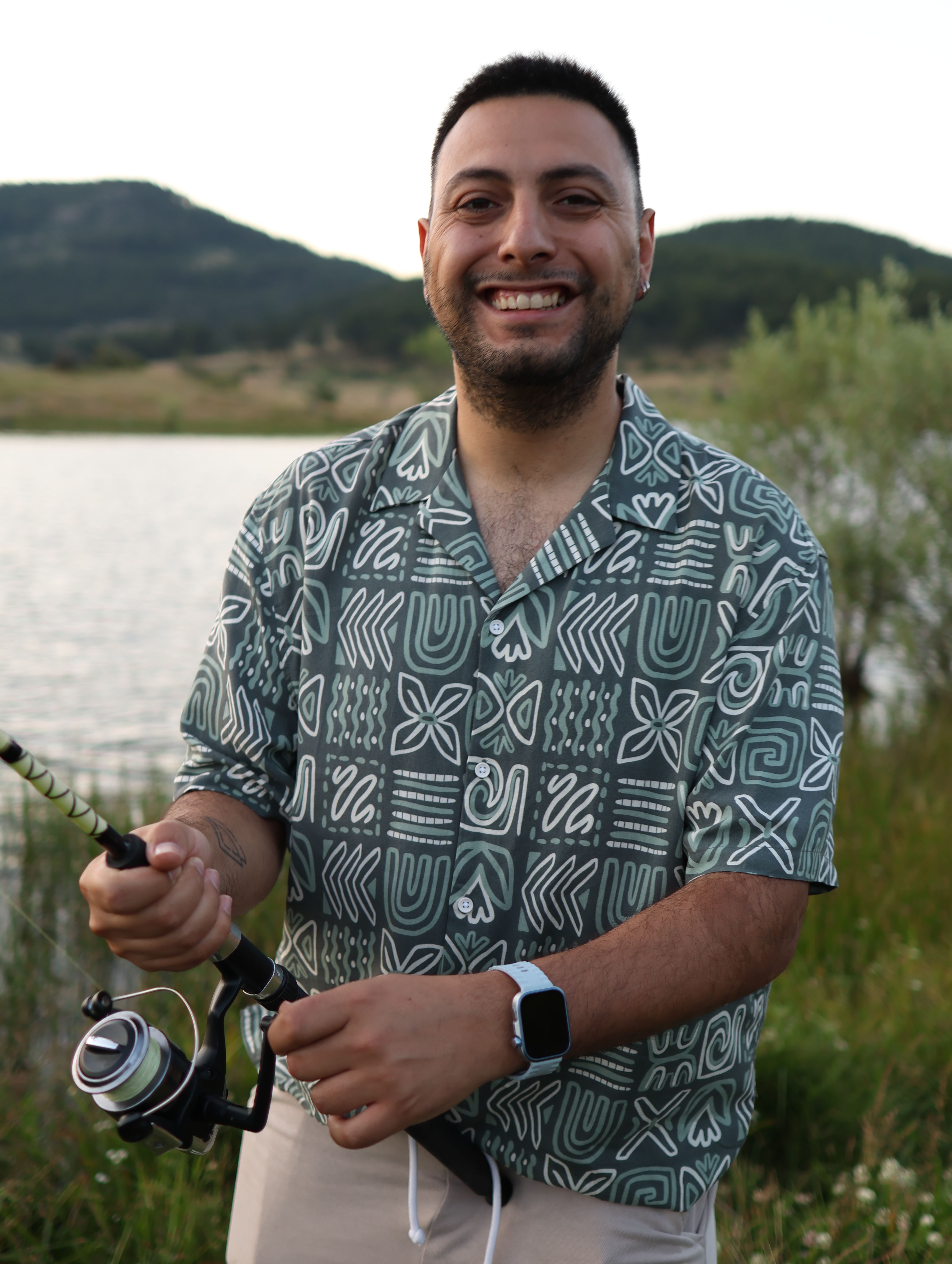 Smiling man holds fishing rod near peaceful lake with mountains in the background during summer.