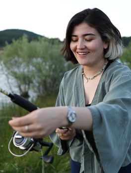 Smiling young woman fishing by a lake during the day, embracing the outdoor experience.