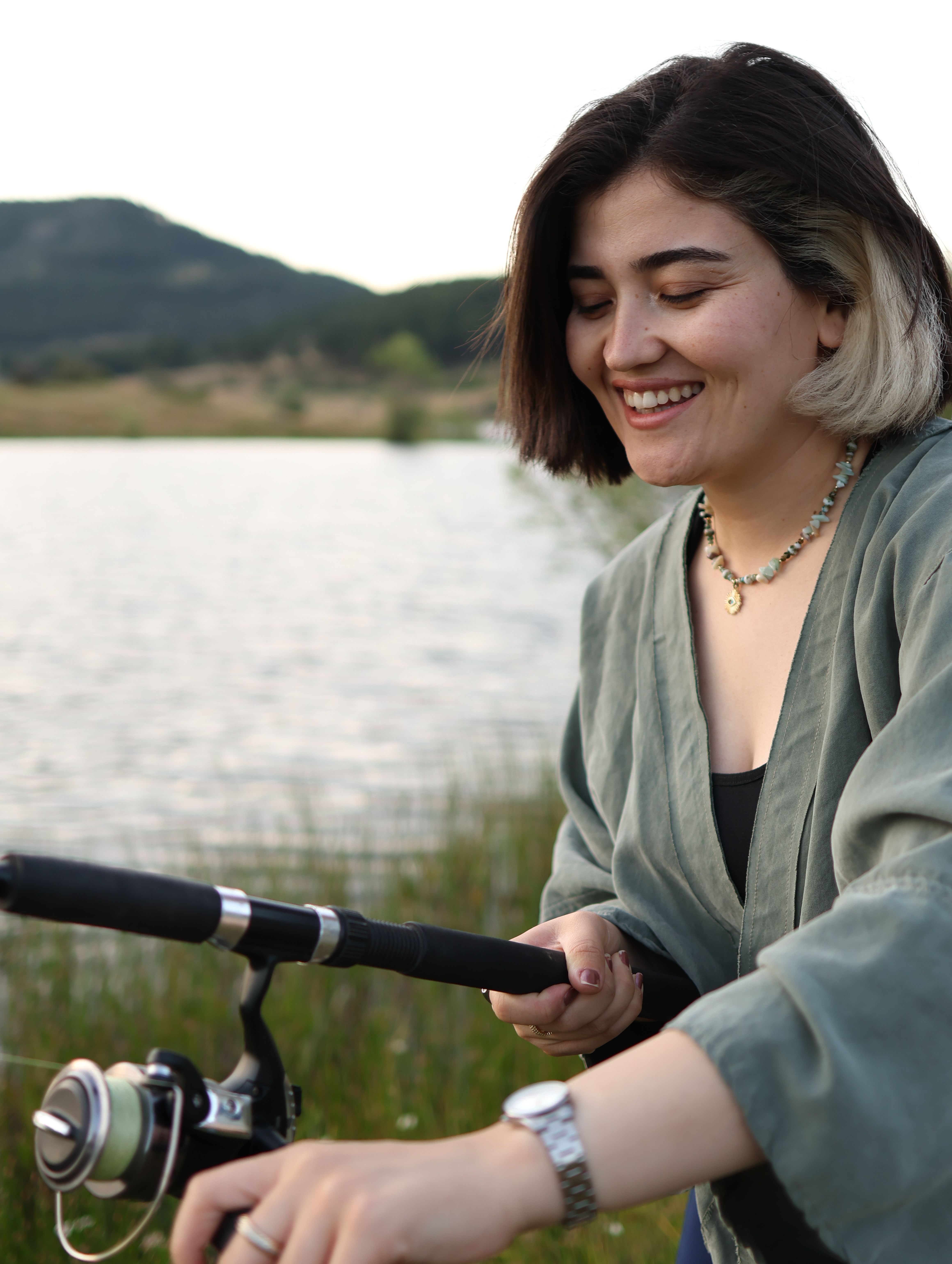 Smiling young woman fishing by a serene lake with lush hills in the background.