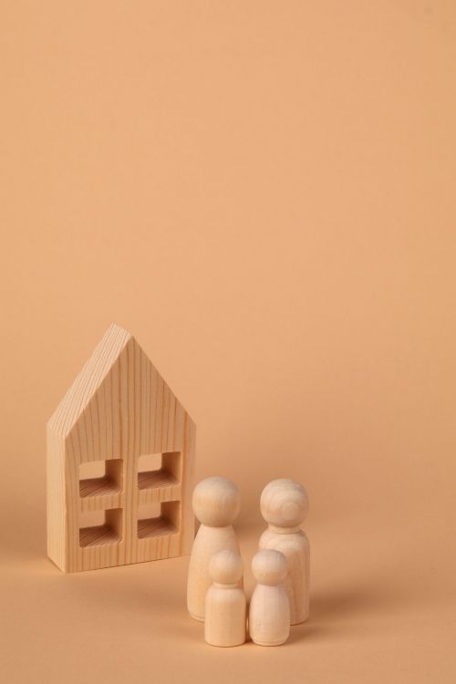 Free Simple wooden pegs represent a family beside a small wooden house model on a beige backdrop. Stock Photo