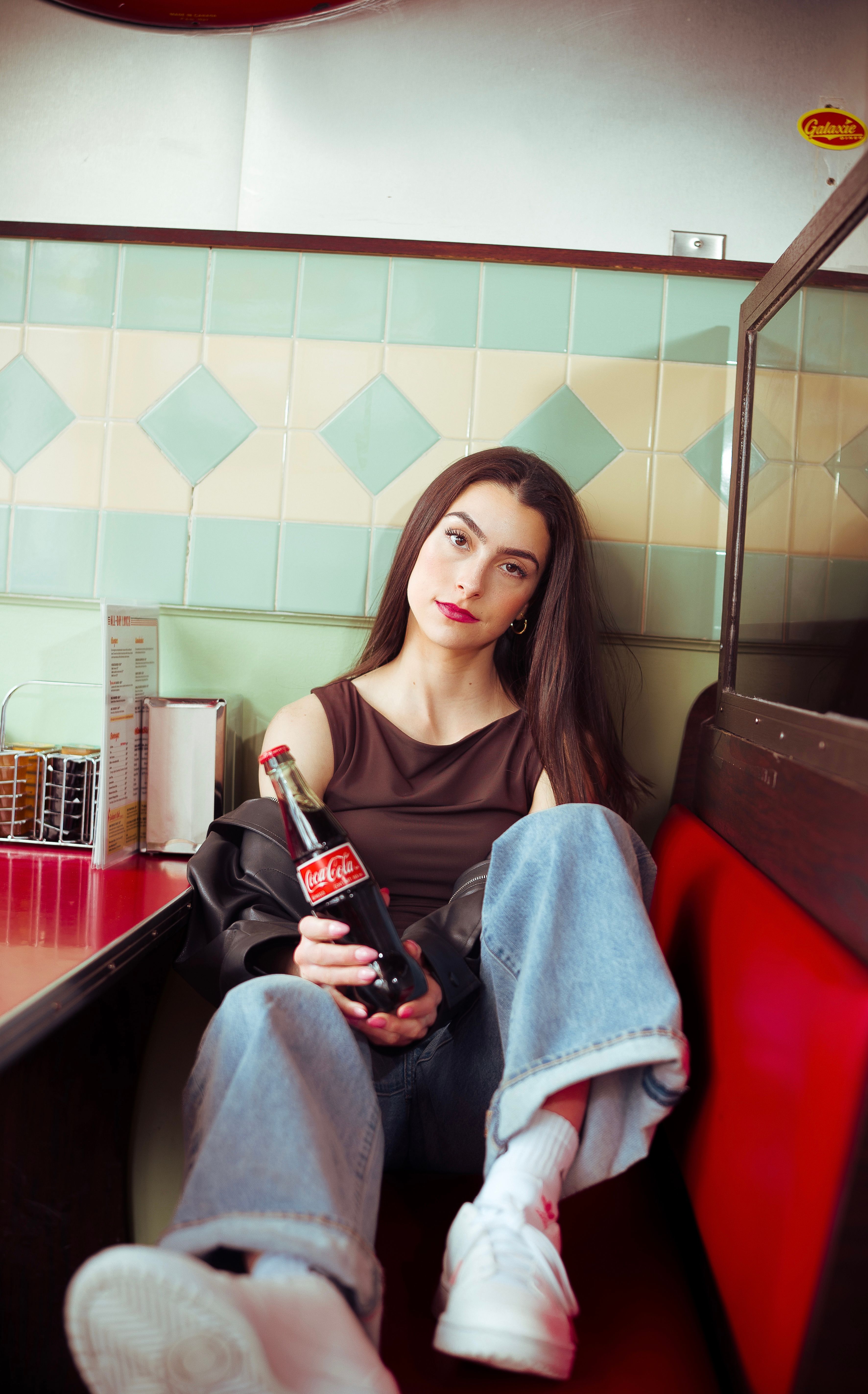 Casual scene of a young woman holding a soda bottle in a vintage diner setting.