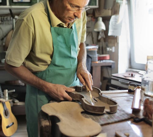 Serious elderly male carpenter in apron and glasses making musical instrument while working on wooden table in workshop