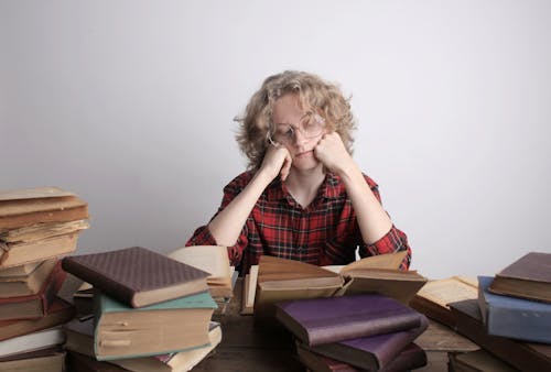 Bored blond haired teenage boy in circle eyeglasses and casual clothes reading book while sitting at table with many various books and propping up head with hands
