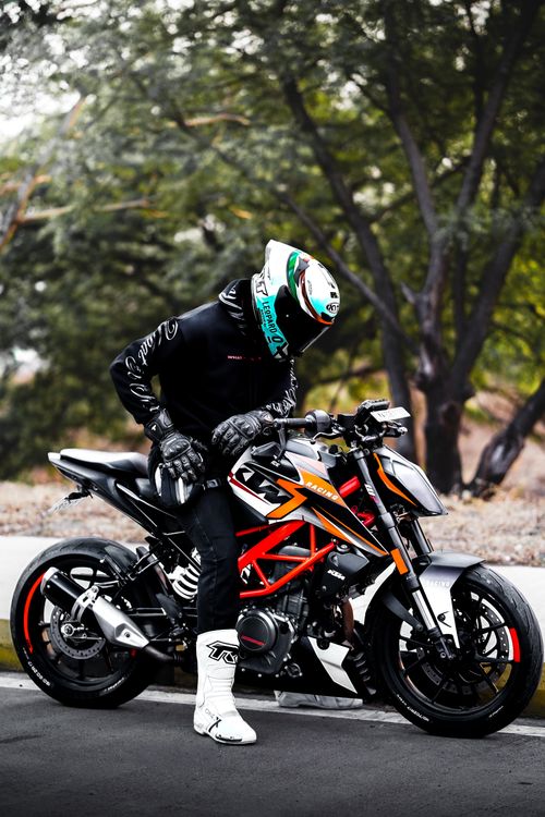 Free A motorcyclist in gear and helmet leaning on a sport bike in a tree-lined outdoor setting. Stock Photo