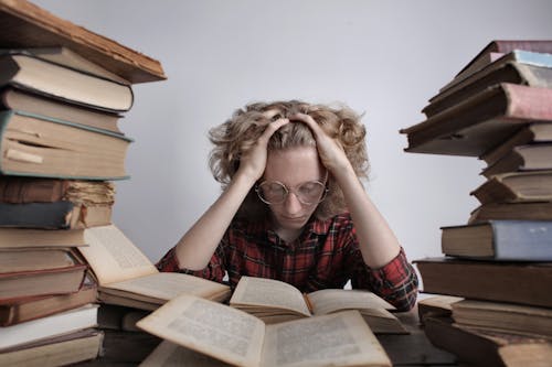 Free Tired teenage boy in casual clothes and eyeglasses sitting at desk with large stacks of books holding head and reading book while preparing for exam Stock Photo
