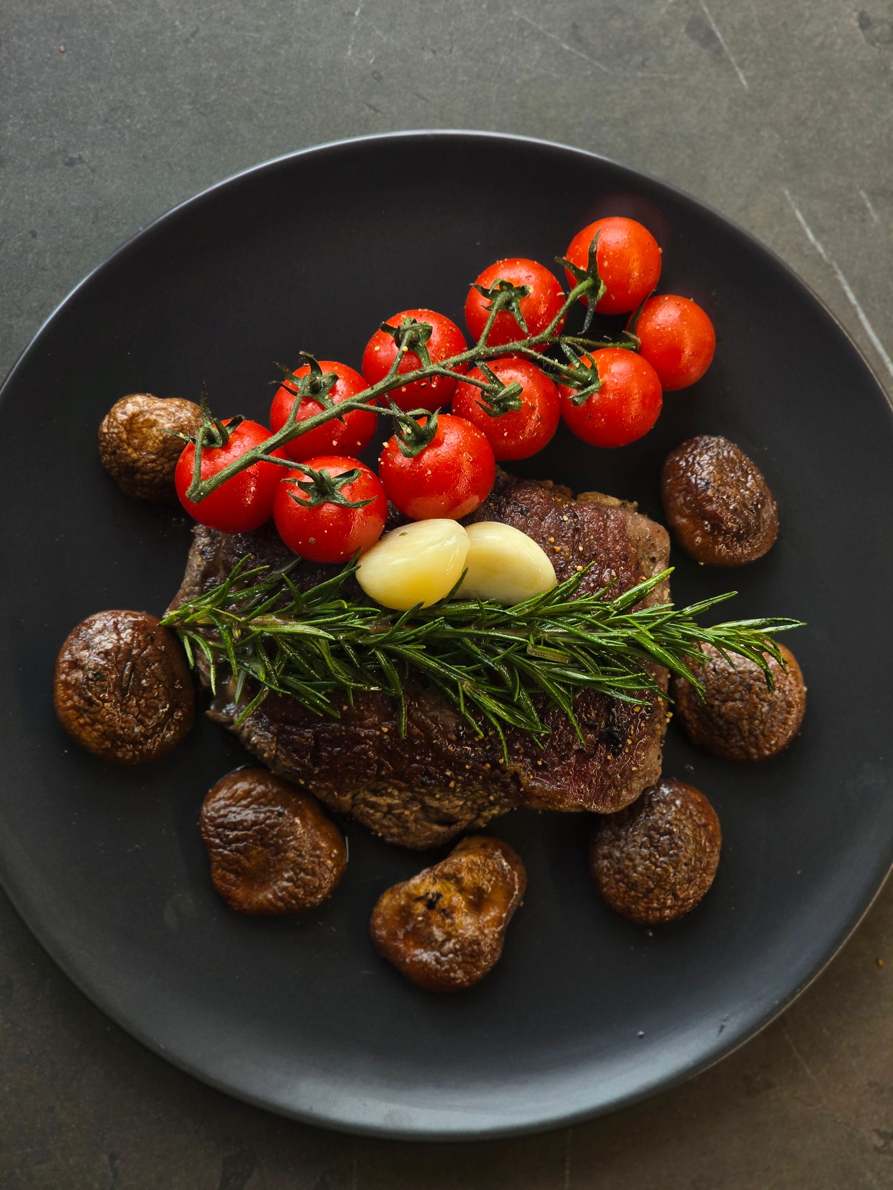 Gourmet steak served with tomatoes, mushrooms, garlic, and rosemary on a black plate.