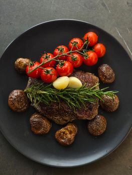 Gourmet steak served with tomatoes, mushrooms, garlic, and rosemary on a black plate.