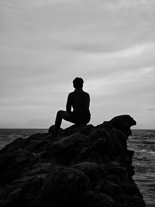 Free A person seated on a rock by the ocean, silhouetted against a cloudy sky, evokes solitude and reflection. Stock Photo