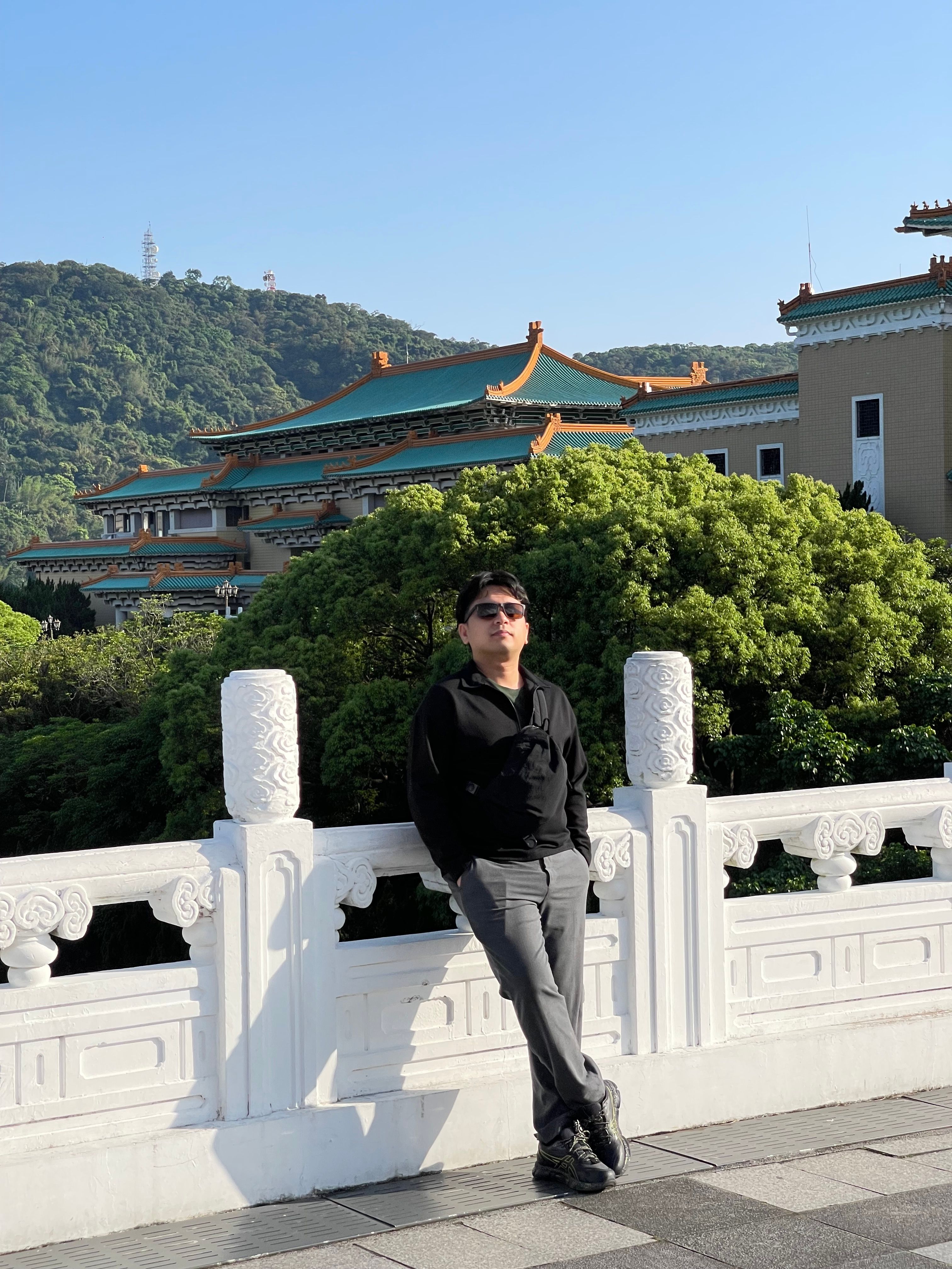 A man in black attire relaxing by the National Palace Museum in Taipei with lush greenery and hills.