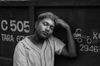 Black and white unhappy thoughtful ethnic male in worn out shirt leaning on metal container and touching forehead while looking at camera with sadness