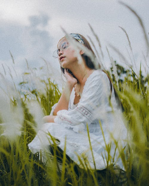 Side view full body romantic Asian lady in white dress sitting on lush grassy lawn with eyes closed and touching neck