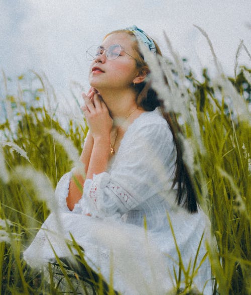 Free Side view full length relaxed young Asian lady in white dress and eyeglasses sitting on lush green meadow with eyes closed while turning face up and touching neck gently Stock Photo