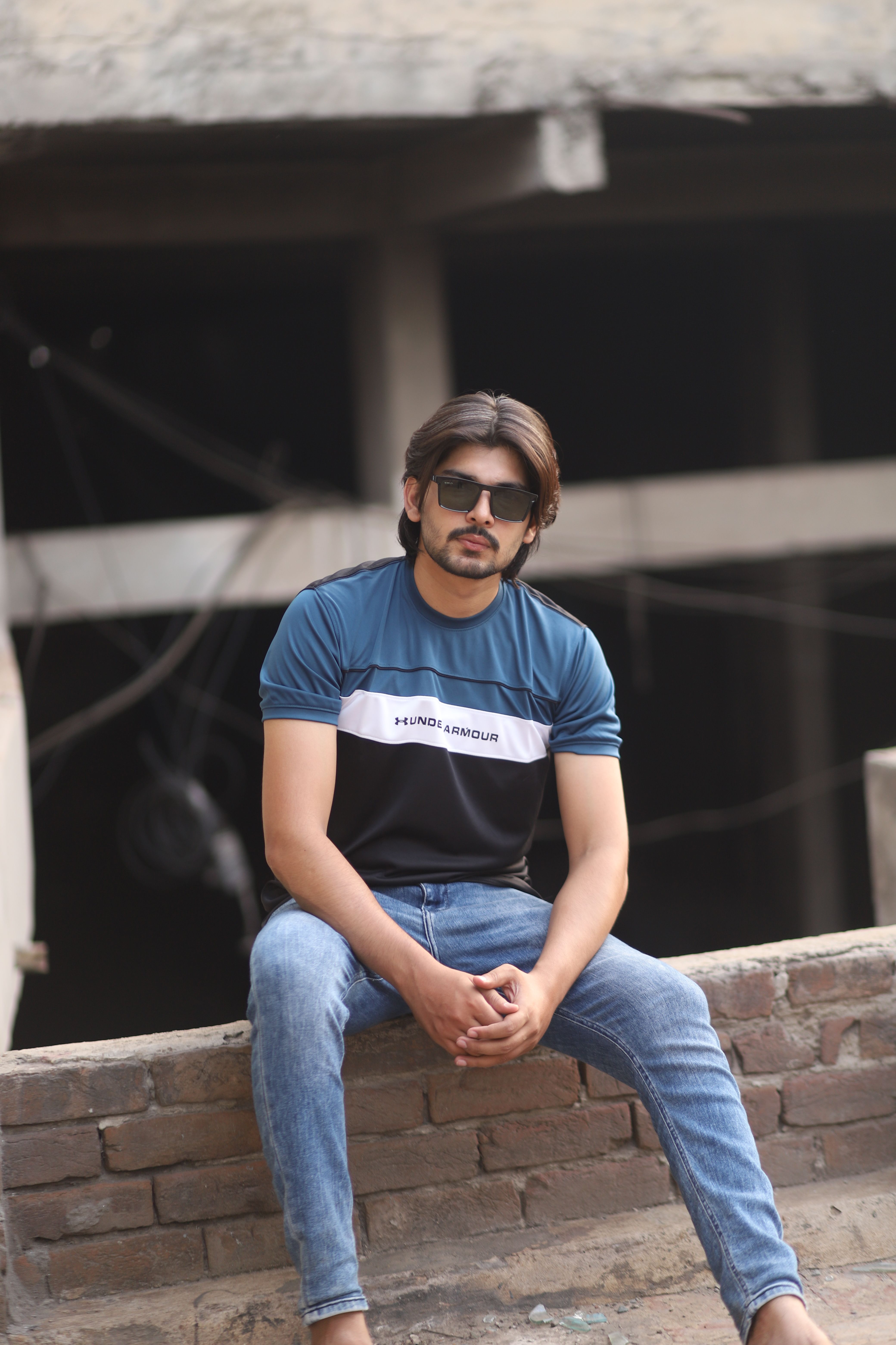 A fashionable young man wearing sunglasses sits on a brick wall in a casual pose.