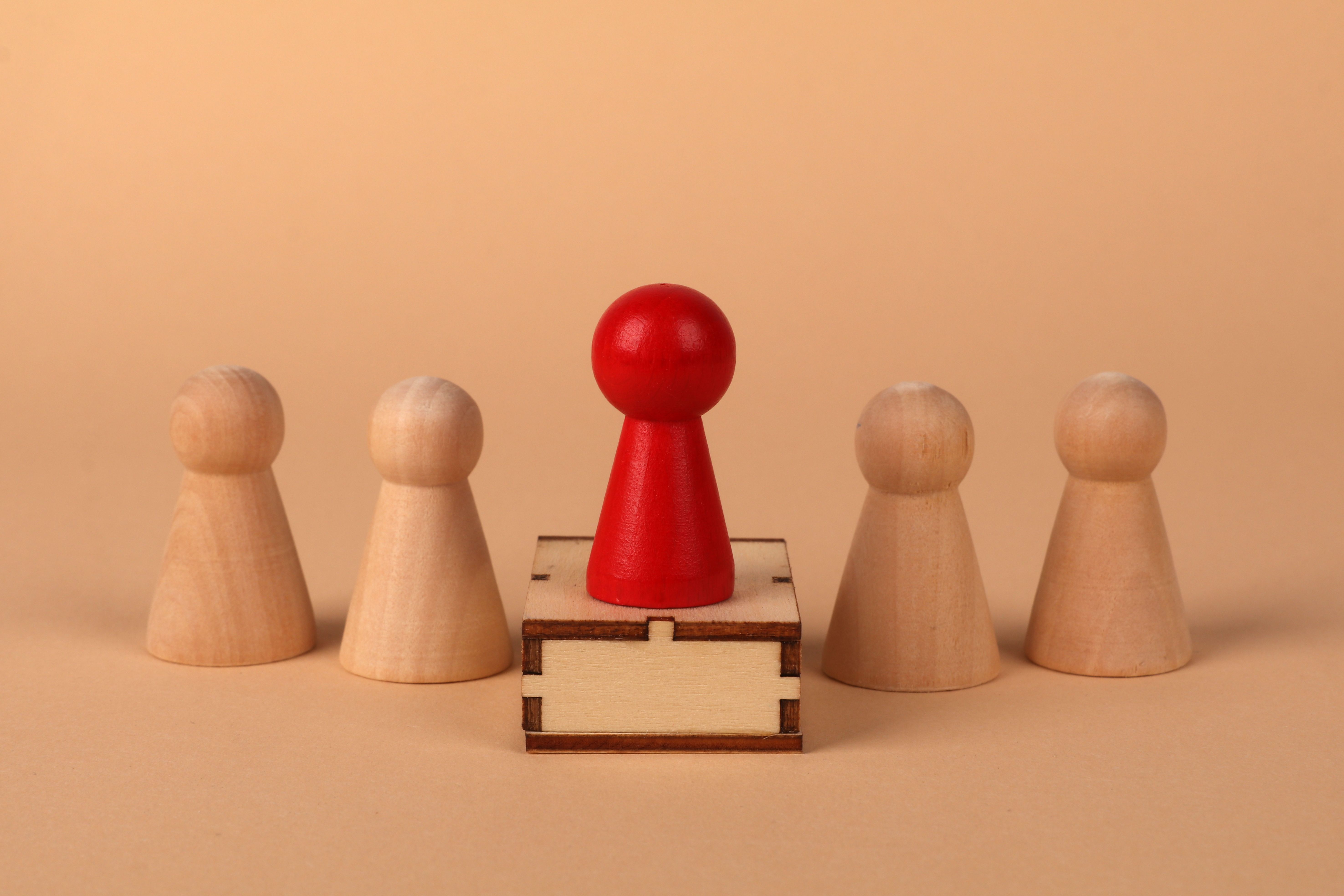 A red pawn stands out as a leader among wooden pieces on a peach background.