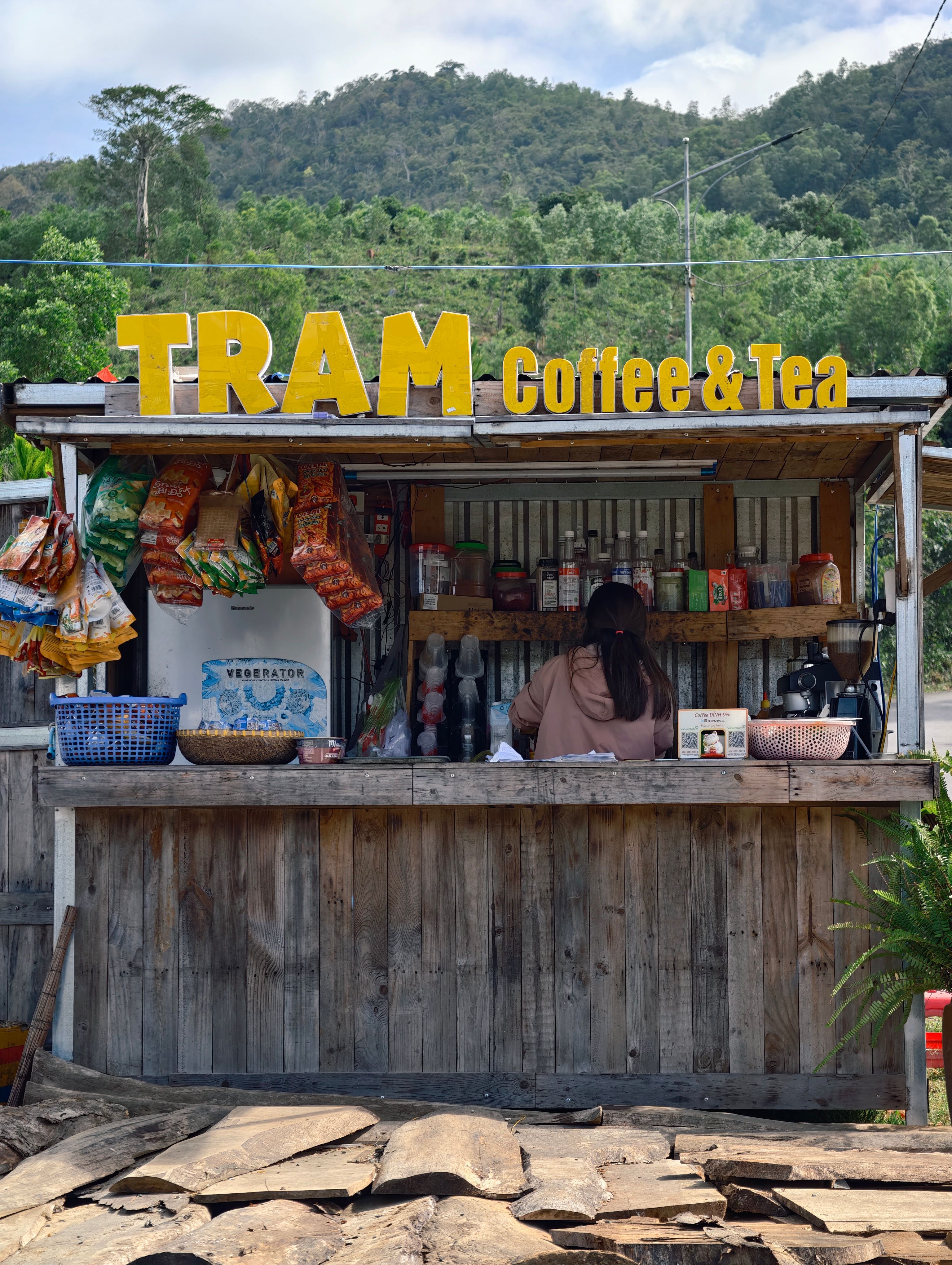 A rustic outdoor coffee and tea stand with snacks, featuring 'Tram' signage, set in a natural landscape.