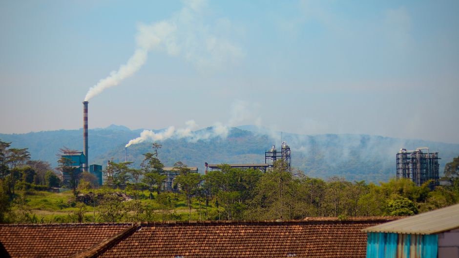 Industrial plant emitting smoke in a lush green area with mountains in the background, indicating pollution
