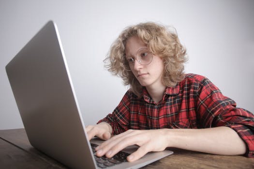 A focused young adult working on a laptop indoors, wearing glasses and a red plaid shirt.