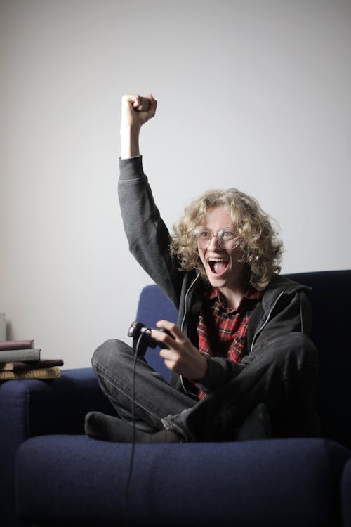 Free Excited youthful guy in casual outfit sitting on sofa with raised hand and rejoicing in victory while playing video game during spending time at home Stock Photo