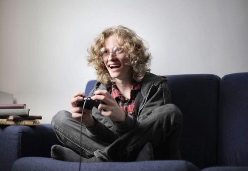 Free Enthusiastic teen boy in casual wear and eyeglasses sitting on blue sofa with legs crossed and playing video game with joystick at home Stock Photo