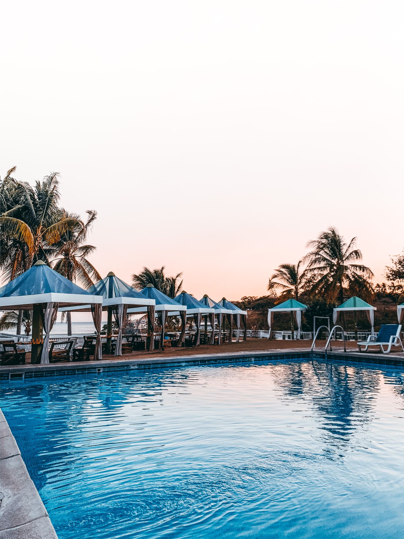 Empty resort swimming pool beside shaded cabanas and palm trees