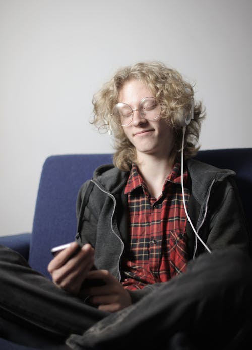 Satisfied teenager with long blond hair in casual outfit and eyeglasses taking pleasure in music on headphones with eyes closed while chilling on sofa with smartphone