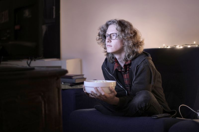 Concentrated male teenage with long hair in casual wear and eyeglasses sitting on couch with bowl of popcorn and watching interesting movie while resting in cozy living room at home
