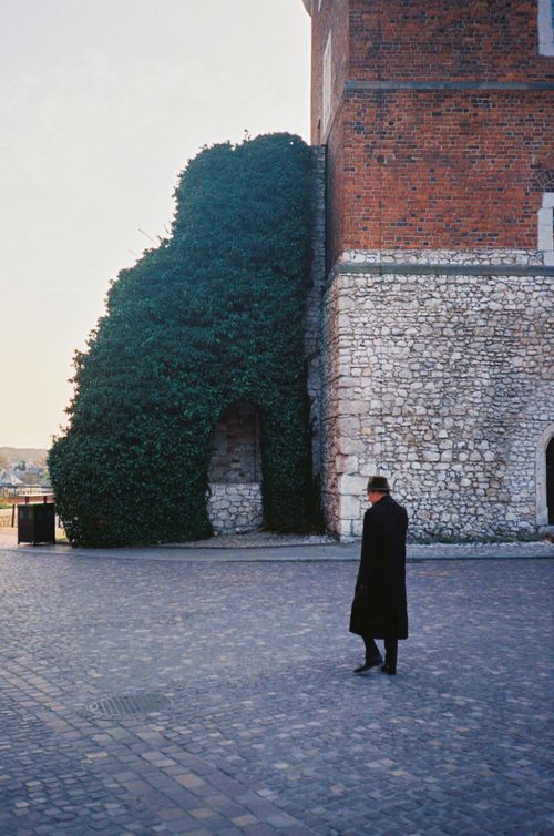 Free A man in a black coat walks alongside an ivy-covered stone building, creating a historic and contemplative scene. Stock Photo