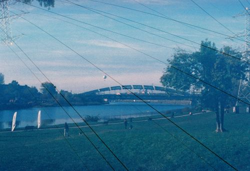 Free A serene view of a riverside park with power lines and a distant bridge under a clear blue sky. Stock Photo