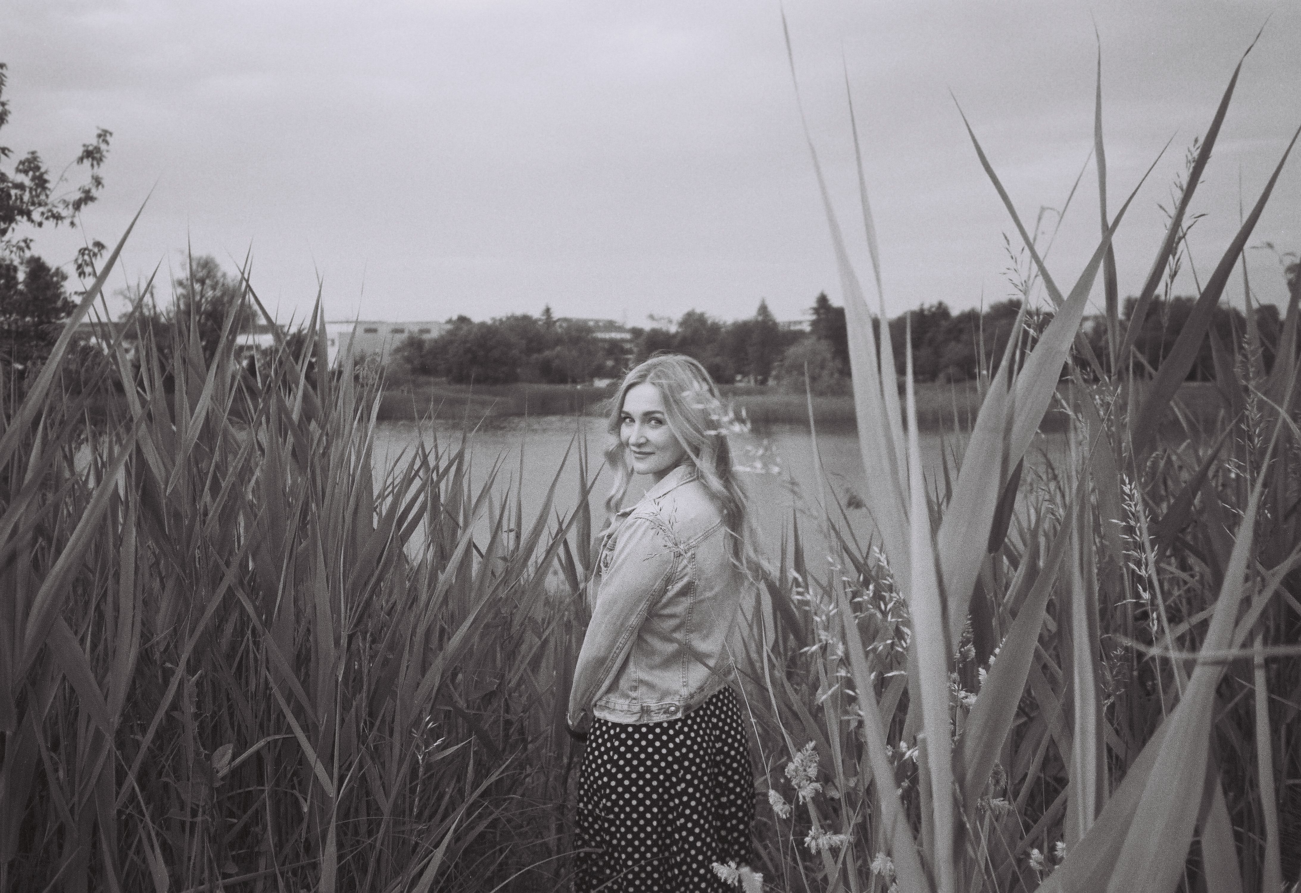 A woman in a denim jacket stands among tall grasses by a lake in monochrome.