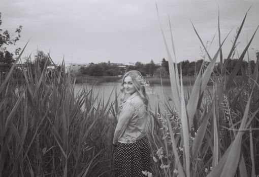 A woman in a denim jacket stands among tall grasses by a lake in monochrome.