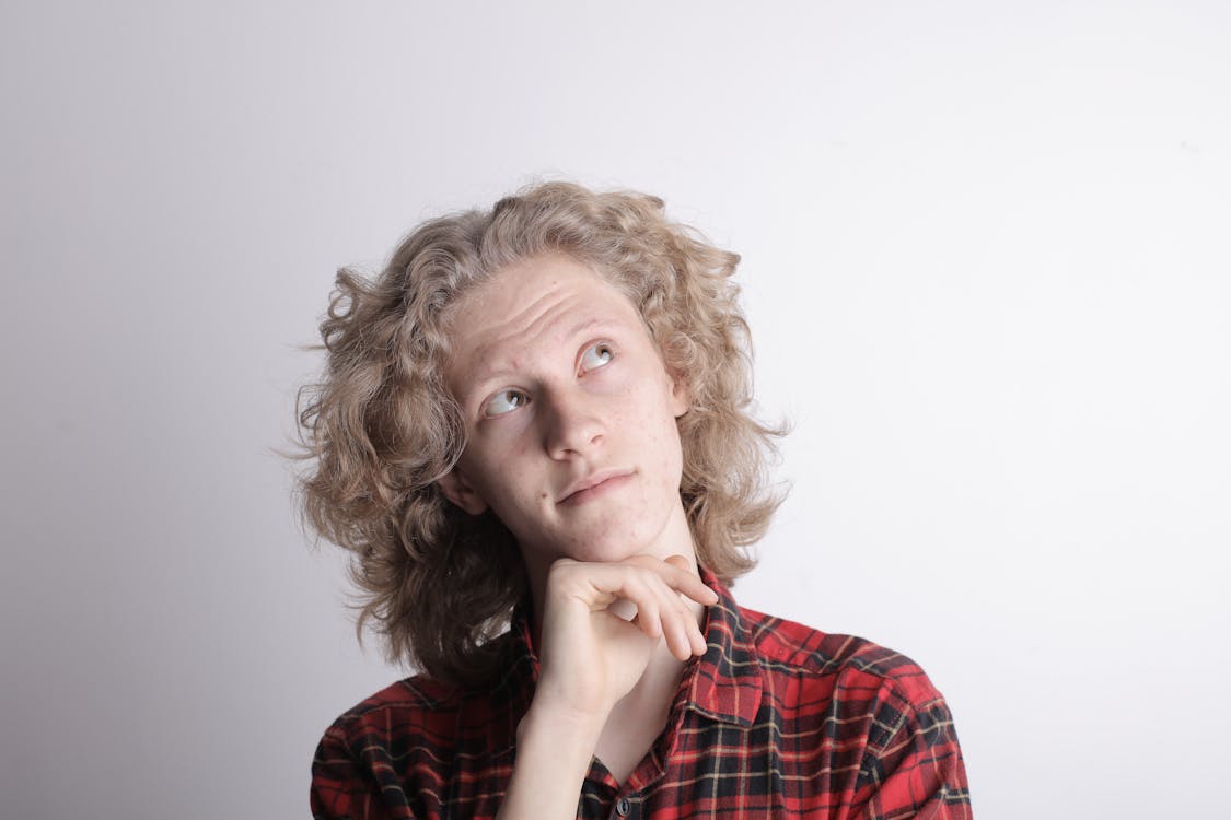 Thoughtful casual teenage boy in checkered red shirt looking up wrinkling forehead and holding chin with hand while standing against gray background