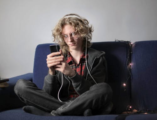 Long haired teenager in circle eyeglasses and casual wear sitting on blue soft couch with luminous garland messaging on mobile phone and listening to music on headphones at home